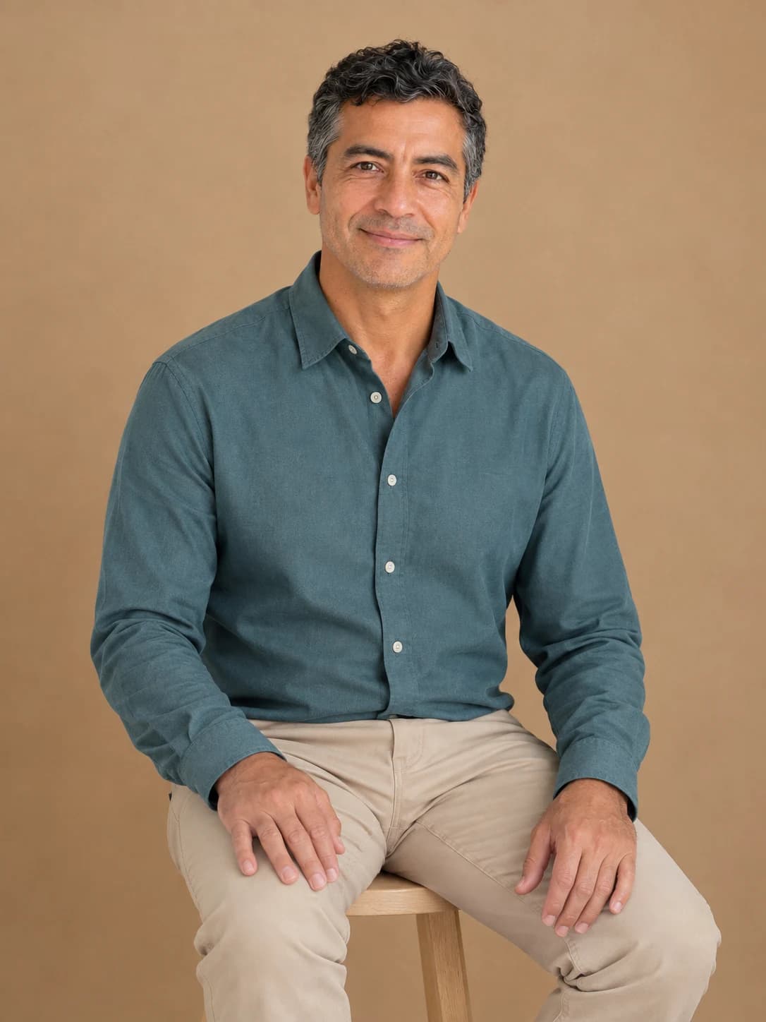 Portret așezat al aceluiași personaj fictiv, în cămașă turcoaz stins și pantaloni bej nisipiu, pe un taburet din lemn deschis, cu fiecare mână relaxată separat pe propria coapsă, într-un studio cu fundal cafeniu cald.